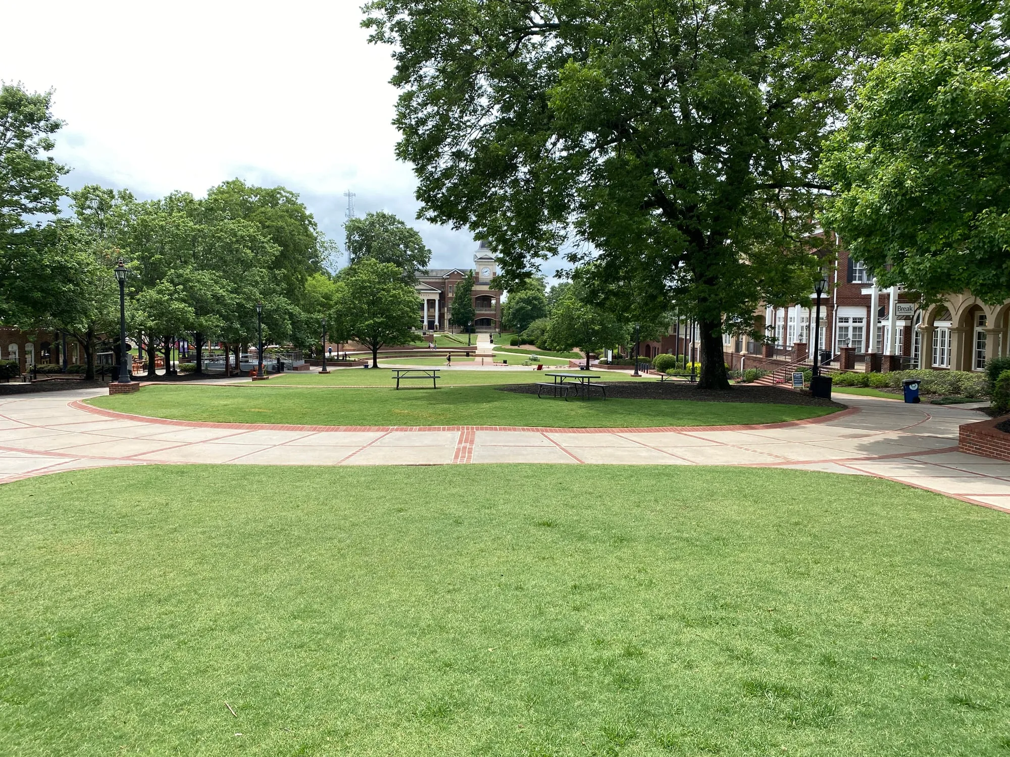 A wide lawn with picnic tables and curving paths in downtown Duluth, framed by shade trees, with a red-brick building with white columns and a white clock tower at the far end.